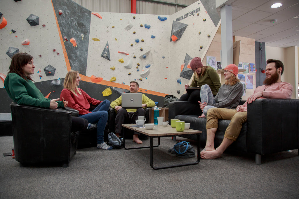 A group of friends having a meeting and a coffee, relaxing in a climbing centre. They are sat on comfy chairs with the climbing wall behind them