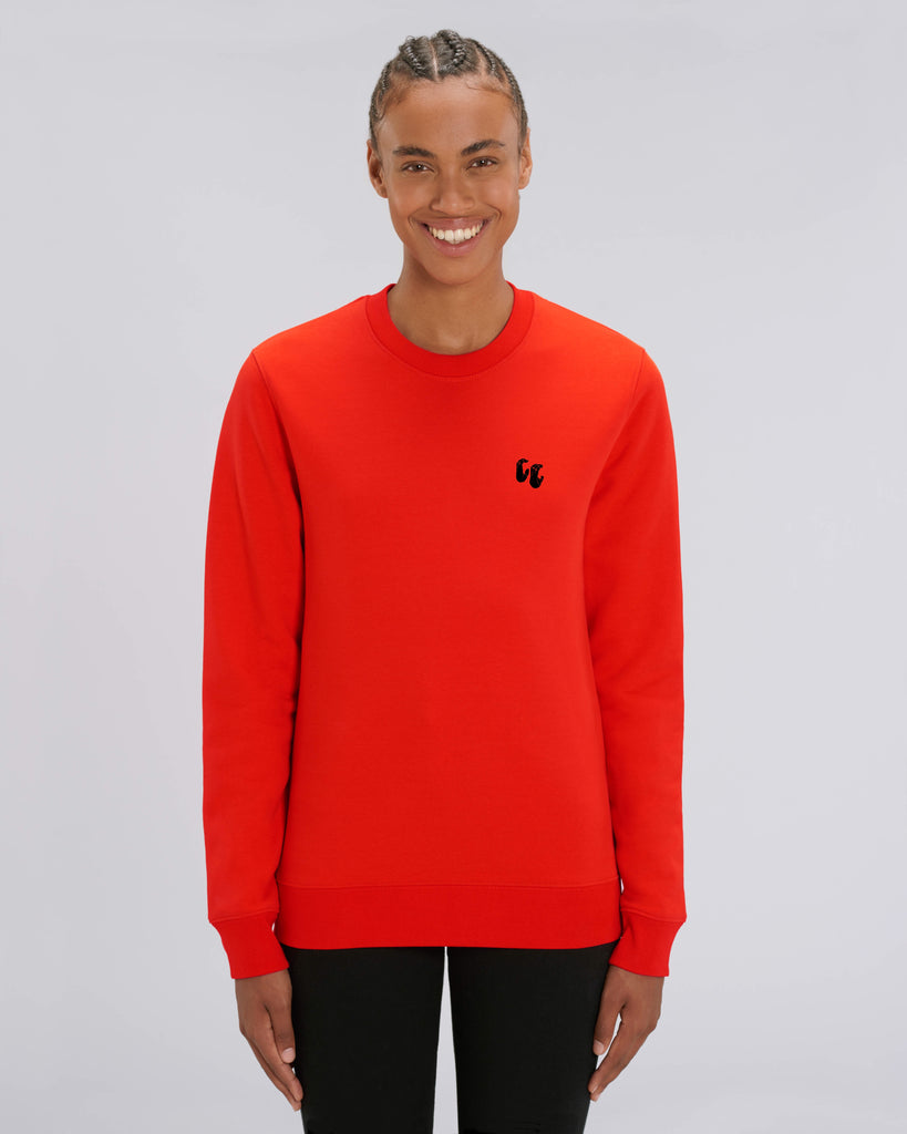 A woman wearing a bright red unisex crew neck sweater, made of organic cotton and recycled polyester, styled with black trousers. The sweater has a small black chest logo in the shape of two hands in a 'crimp' climbing grip position