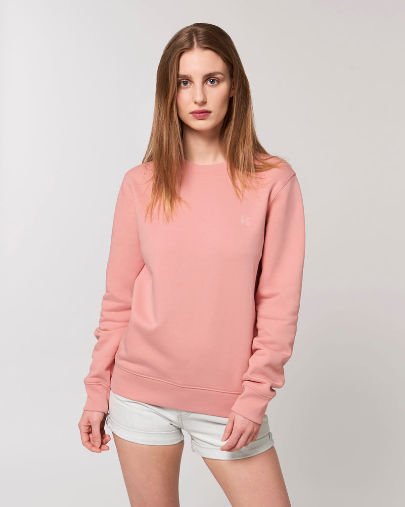 A woman wearing a dusky pink unisex crew neck sweater, made of organic cotton and recycled polyester, styled with white shorts. The sweater has a small pale pink chest logo in the shape of two hands in a 'crimp' climbing grip position