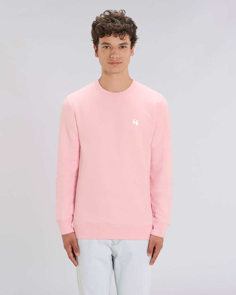 A man wearing a cotton pink unisex crew neck sweater, made of organic cotton and recycled polyester, styled with pale jeans. The sweater has a small white chest logo in the shape of two hands in a 'crimp' climbing grip position