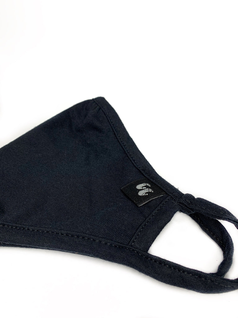 A black face mask with fabric loops for the ears. Made of Organic cotton and recycled polyester it has a subtle fabric logo label stitched to the left hand side