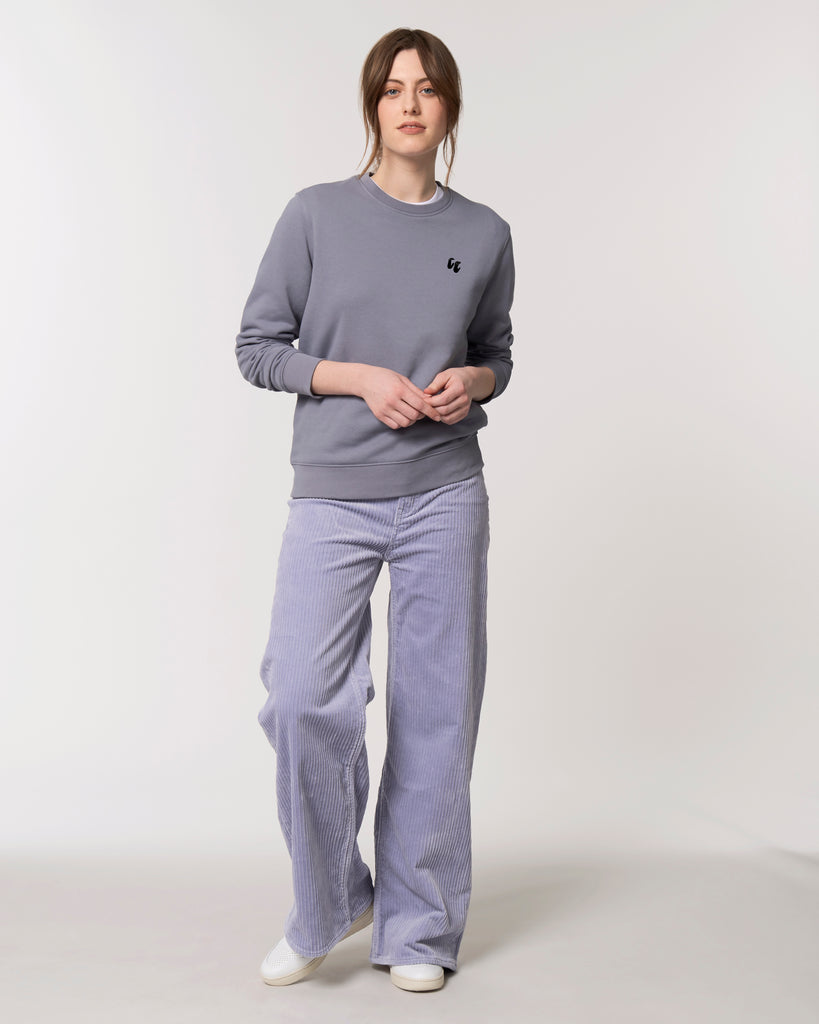 A woman wearing a blue grey unisex crew neck sweater, made of organic cotton and recycled polyester, styled with lilac cords. The sweater has a small black chest logo in the shape of two hands in a 'crimp' climbing grip position