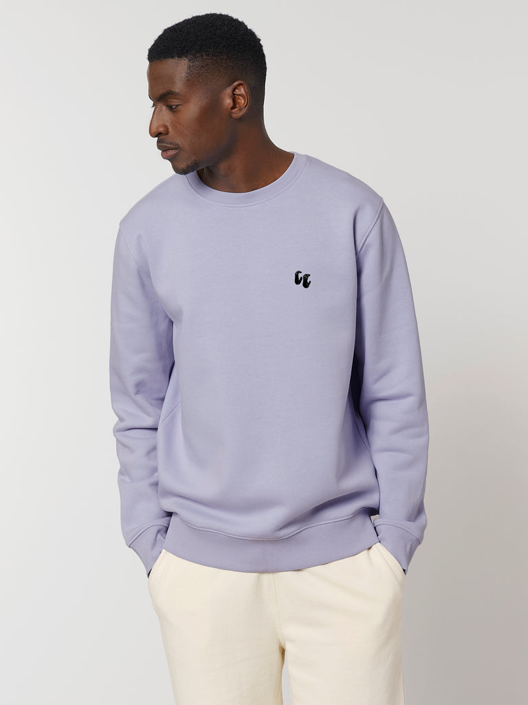 A man wearing a lavender purple unisex crew neck sweater, made of organic cotton and recycled polyester, styled with white trousers. The sweater has a small black chest logo in the shape of two hands in a 'crimp' climbing grip position