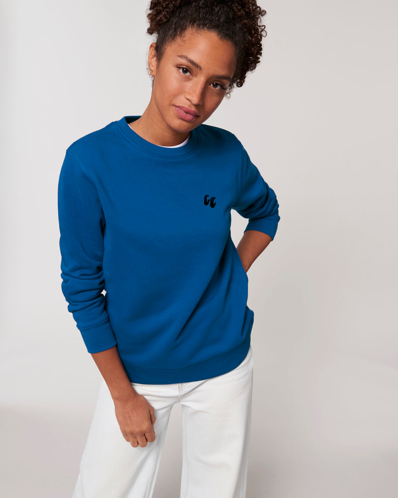 A woman wearing a bright blue unisex crew neck sweater, made of organic cotton and recycled polyester, styled with white trousers. The sweater has a small black chest logo in the shape of two hands in a 'crimp' climbing grip position