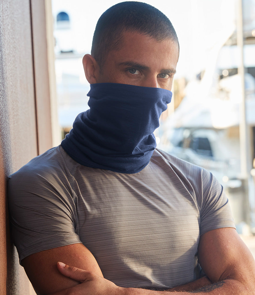A man stood casually leaning against a wall wears a neutral, striped t-shirt and a Navy Blue Merino Wool Neck Gaiter
