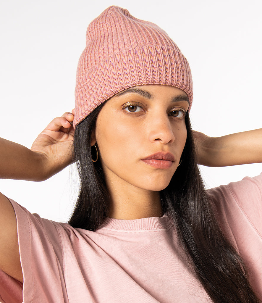 A close up of a woman with long, dark hair wearing a soft pink, ribbed, Crimpd Clothing beanie made from 100% merino wool