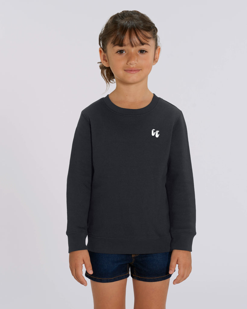 A young girl wearing a black crew neck sweater, styled with blue jean shorts. The sweater has a small white logo on the left chest position which is of two hands in a crimp climbing position
