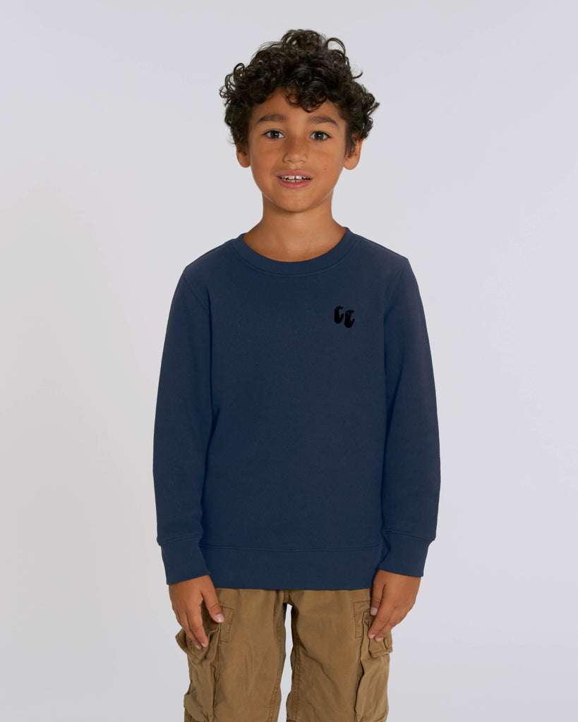 A young boy wearing a navy blue crew neck sweater, styled with beige trousers. The sweater has a small black logo on the left chest position which is of two hands in a crimp climbing position