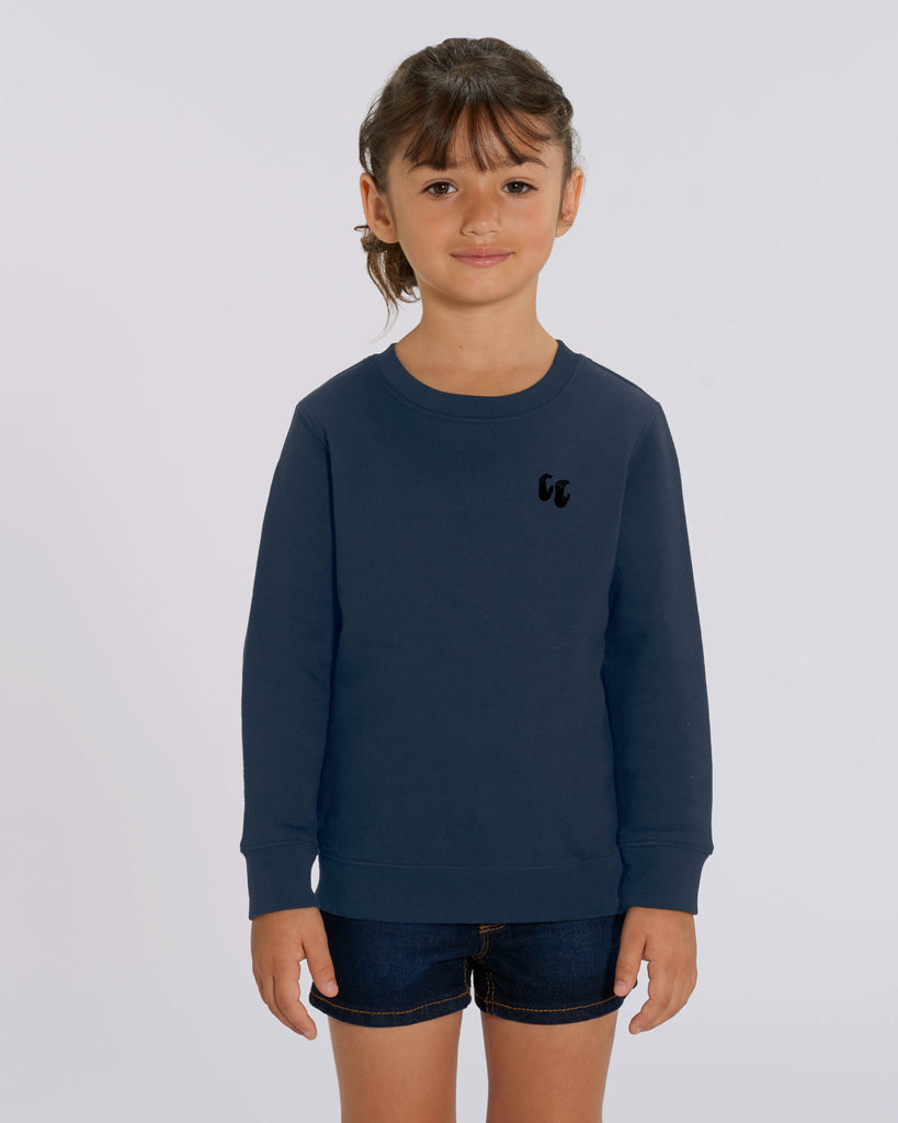 A young girl wearing a navy blue crew neck sweater, styled with denim shorts. The sweater has a small black logo on the left chest position which is of two hands in a crimp climbing position