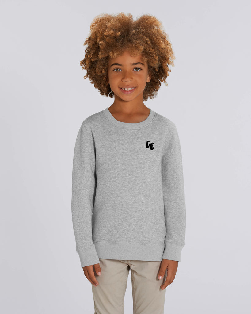 A young boy with curly hair wearing a light heather grey crew neck sweater, styled with beige trousers. The sweater has a small black logo on the left chest position which is of two hands in a crimp climbing position