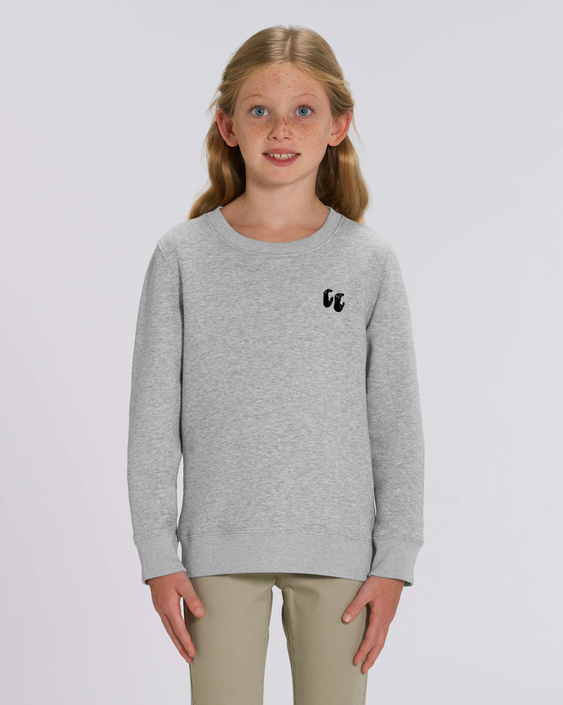 A young girl with curly hair wearing a light heather grey crew neck sweater, styled with beige trousers. The sweater has a small black logo on the left chest position which is of two hands in a crimp climbing position