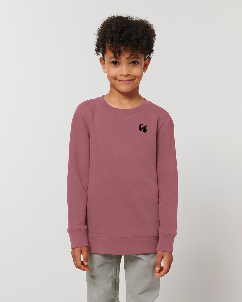 A smiling child wearing a dusky rose crew neck sweater, styled with grey trousers. The sweater has a small black logo on the left chest position which is of two hands in a crimp climbing position