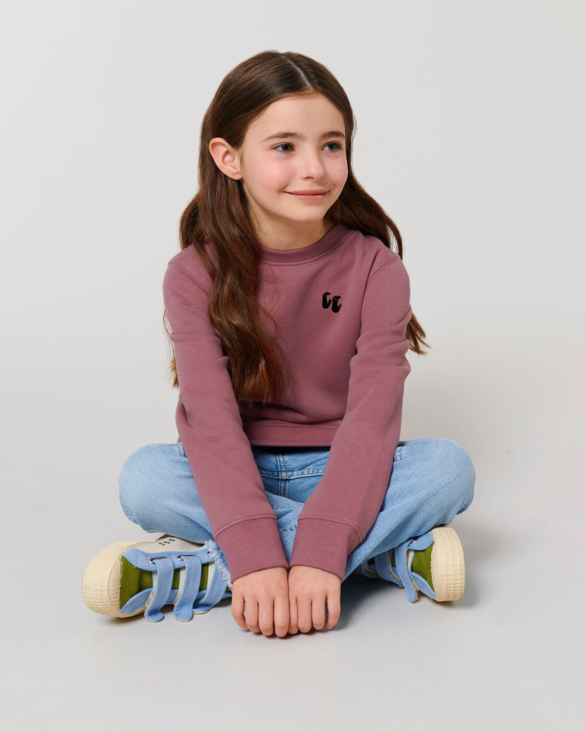 A young girl sat cross-legged wearing a dusky rose sweater made from organic cotton. The sweater has a small black logo on the left chest position and is worn with blue jeans and trainers