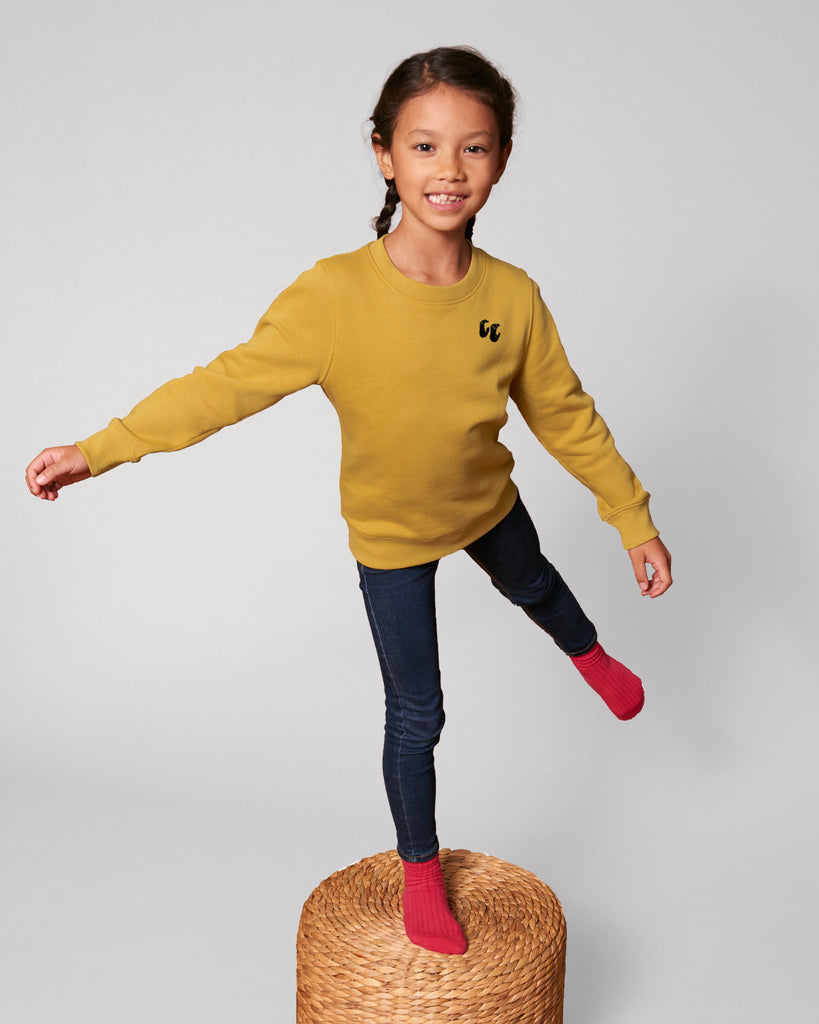 A smiling child with plaits standing on one leg on a woven stool, wearing a mustard yellow crew neck. The jumper is styled with blue jeans and pink socks and has a small black logo on the left chest position