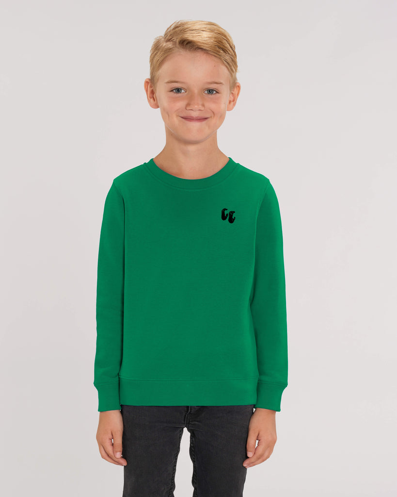 A smiling boy wearing a bright green crew neck sweater, styled with black trousers. The sweater has a small black logo on the left chest position which is of two hands in a crimp climbing position