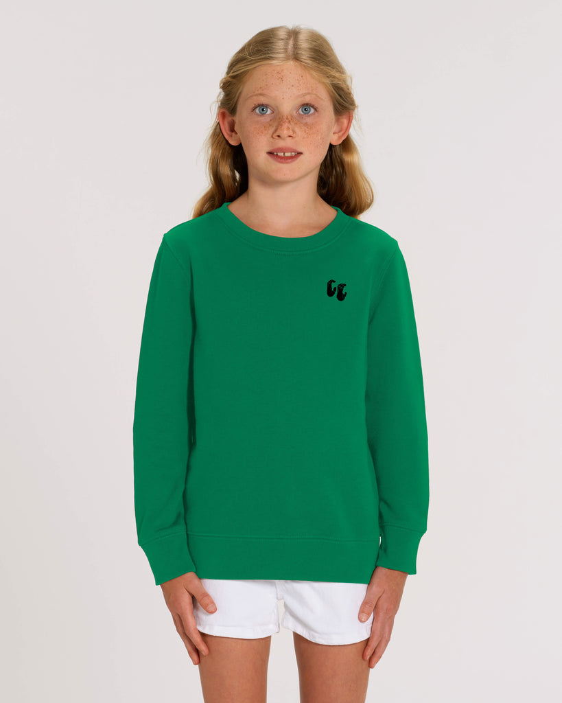 A young girl wearing a bright green crew neck sweater, styled with white shorts. The sweater has a small black logo on the left chest position which is of two hands in a crimp climbing position