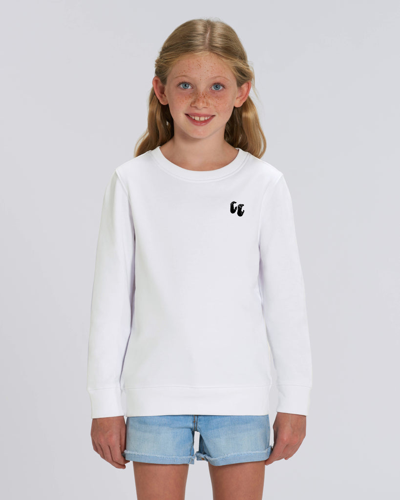 A young girl wearing a white crew-neck sweater, styled with denim shorts. The sweater has a small black logo on the left chest position which is of two hands in a crimp climbing position