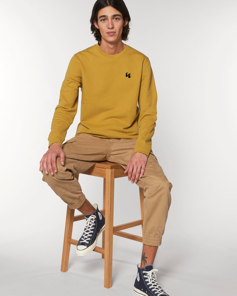A man wearing a mustard yellow unisex crew neck sweater, made of organic cotton and recycled polyester, styled with smart trousers. The sweater has a small black chest logo in the shape of two hands in a 'crimp' climbing grip position