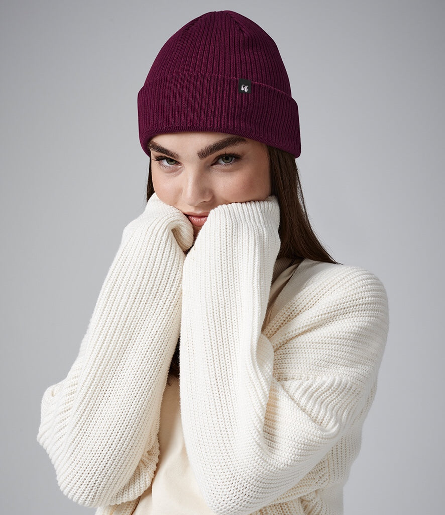 A person wearing a thick, warm, burgundy beanie hat made of organic cotton and has the cuff folded up. There is a fabric label folded over the top of the cuff positioned slightly to the left. Worn with a cream, long sleeved cosy jumper. 