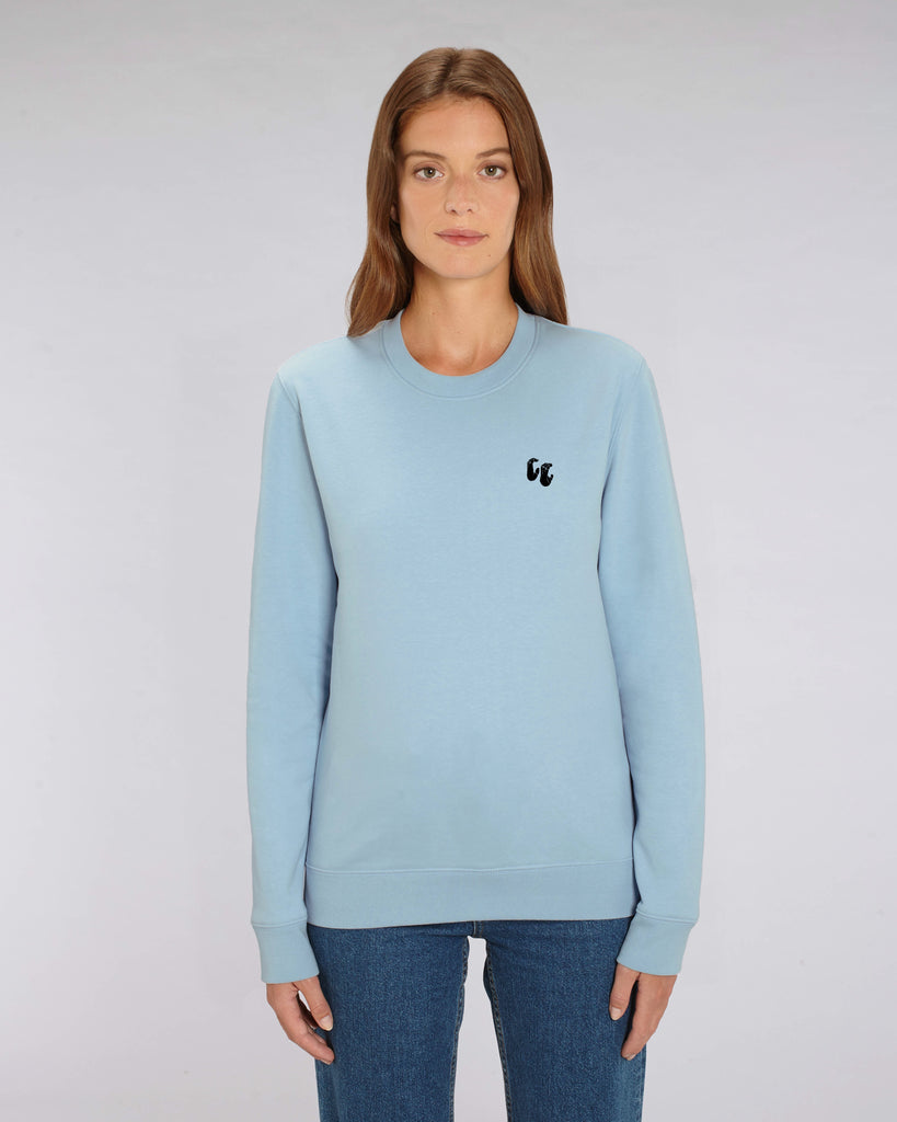 A woman wearing a sky blue unisex crew neck sweater, made of organic cotton and recycled polyester, styled with blue jeans. The sweater has a small black chest logo in the shape of two hands in a 'crimp' climbing grip position