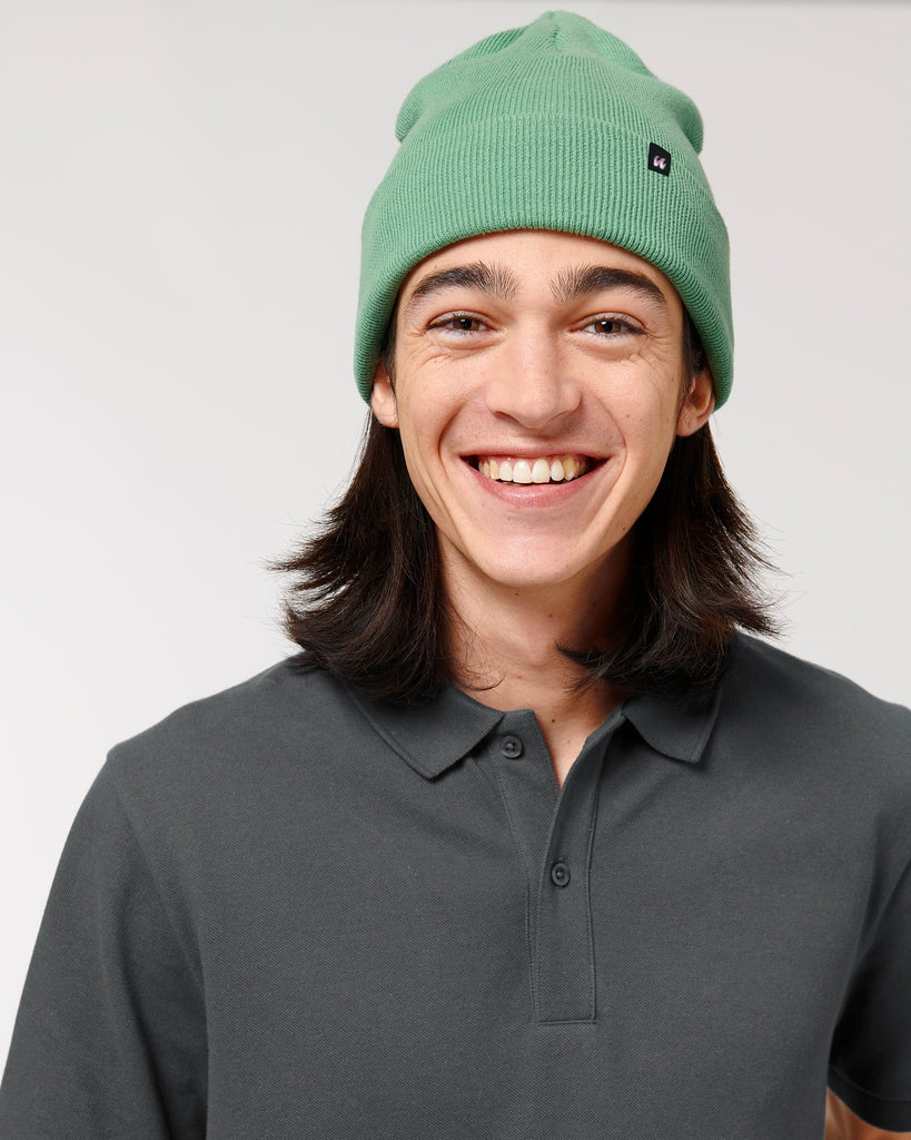 A person wearing a dusty mint green beanie hat that has a small black fabric label stitched to the folded cuff