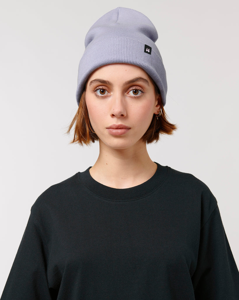 A person wearing a pastel lavender purple beanie hat that has a small black fabric label stitched to the folded cuff
