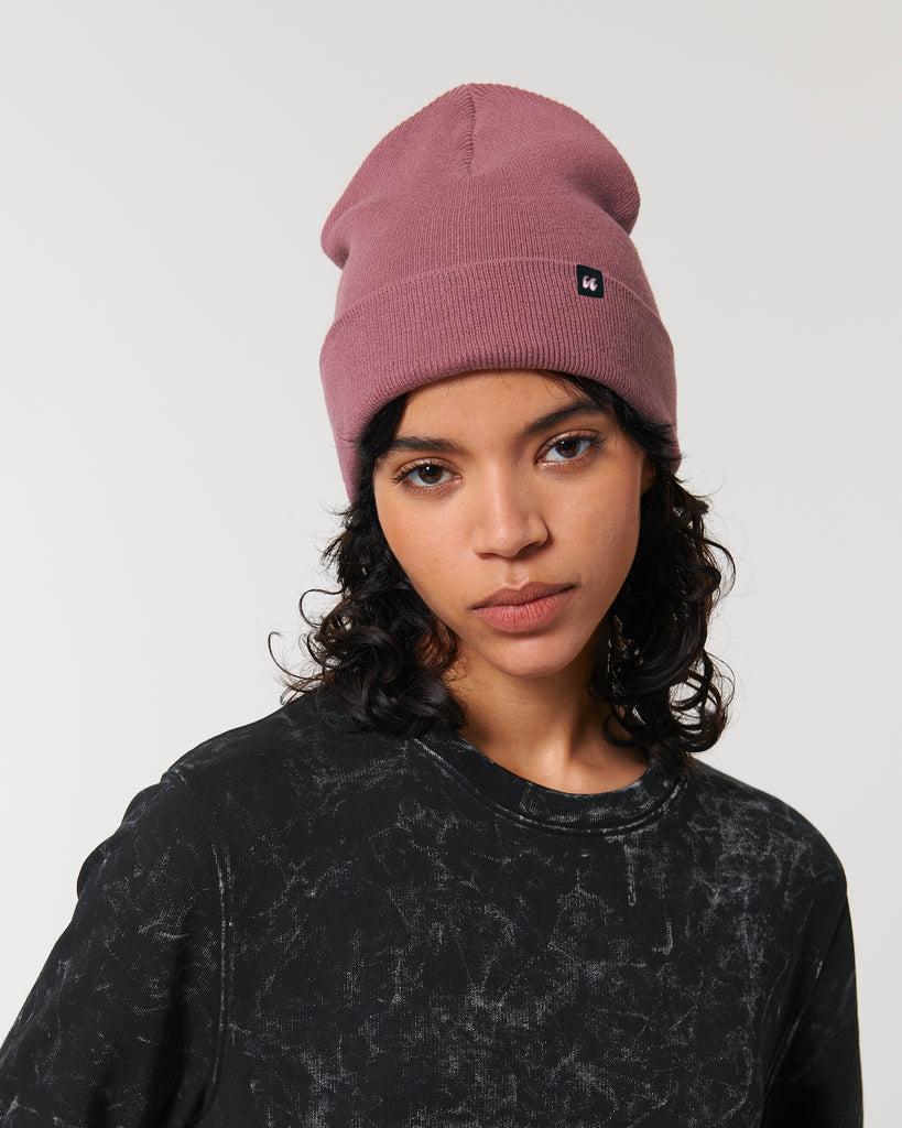 A person wearing a dusky rose pink beanie hat that has a small black fabric label stitched to the folded cuff