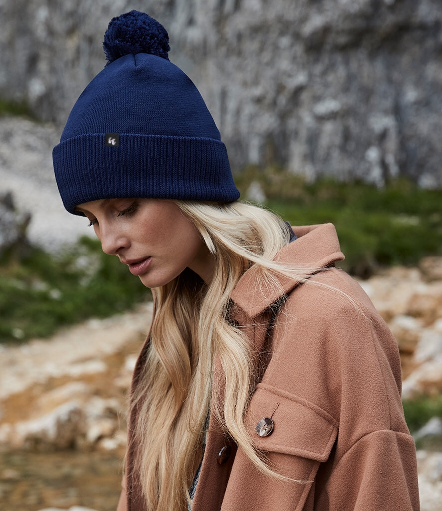 A woman wearing a navy blue bobble hat beanie with pom pom made from organic cotton. She has long blonde hair and has styled it with a camel coat 