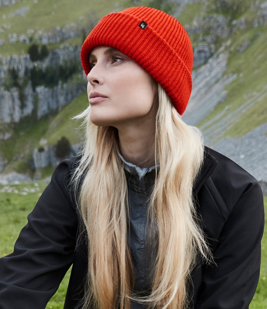 A blonde haired woman wears a recycled polyester beanie in bright red, it has a small black fabric tag on the front left-hand side. 